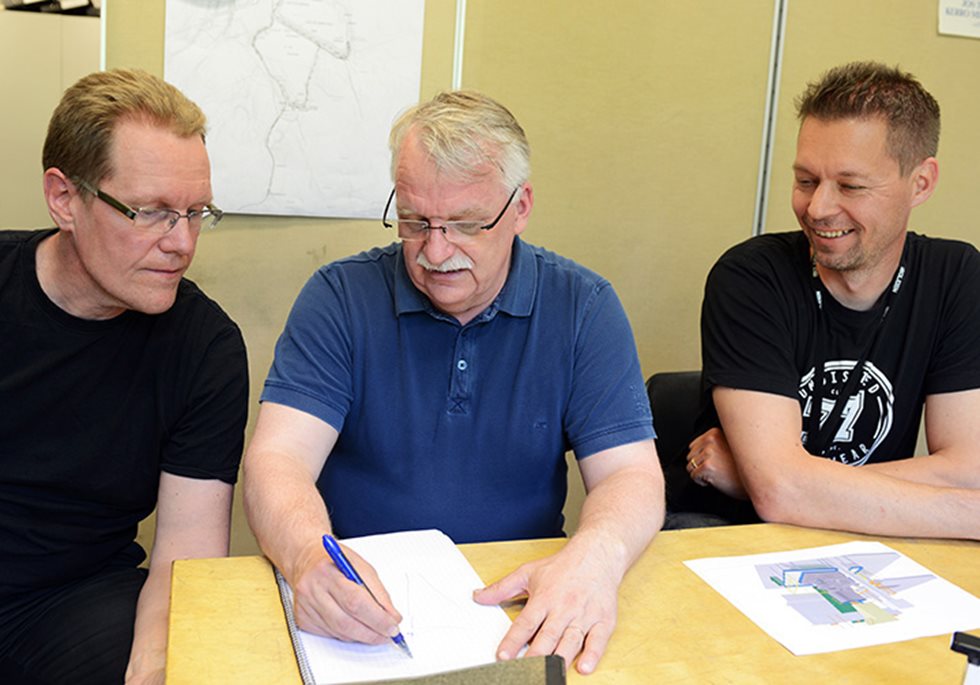 Três homens no escritório. O do meio está escrevendo em um papel enquanto os outros dois estão olhando.