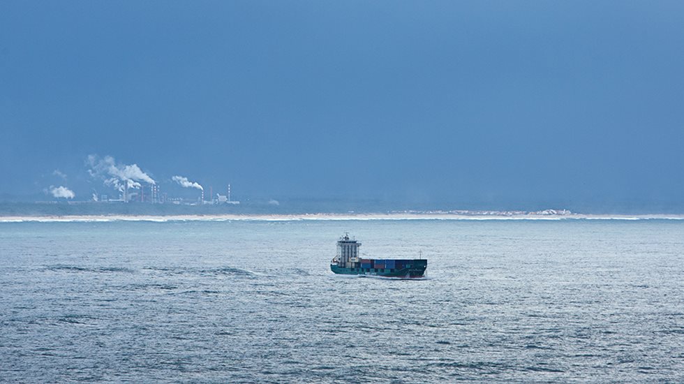 A ship with cargo pictured in the middle of the sea with coast in the far horizon.
