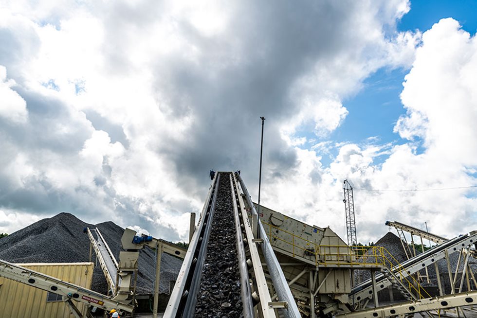 Conveyor belt with aggregate on it pictured against cloudy sky.